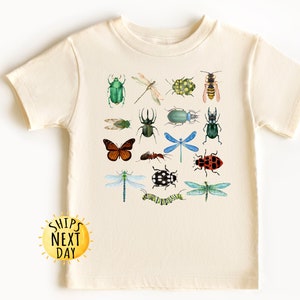 May include: A cream-colored t-shirt with a colorful insect design. The design features a variety of insects, including butterflies, dragonflies, beetles, and ants. The shirt has short sleeves and a crew neck.