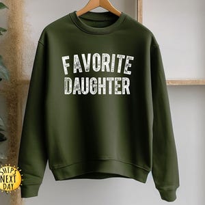 May include: An olive green sweatshirt with the words "FAVOURITE DAUGHTER" printed in white, block letters. The sweatshirt is hanging on a wooden hanger. The image also includes a "SHIPS NEXT DAY" sticker.