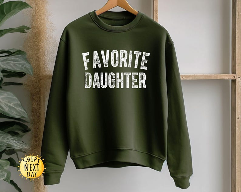 May include: An olive green sweatshirt with the words "FAVOURITE DAUGHTER" printed in white, block letters. The sweatshirt is hanging on a wooden hanger. The image also includes a "SHIPS NEXT DAY" sticker.