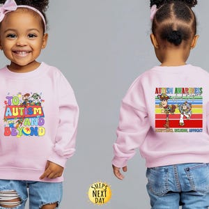 K&ouml;nnte beinhalten: Hellrosa Sweatshirt mit dem Text "TO AUTISM AND BEYOND" in bunten Buchstaben und einer Grafik von Zeichentrickfiguren. Auf der R&uuml;ckseite des Sweatshirts steht "AUTISM AWARENESS And Beyond" mit einem Regenbogenstreifen-Design und den Worten "ACCEPTANCE, INCLUSION, ADVOCACY".