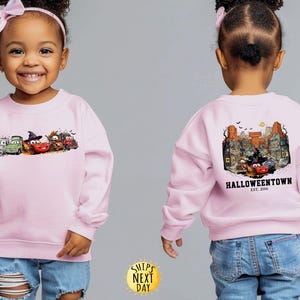 K&ouml;nnte beinhalten: Hellrosa Sweatshirt mit einem Halloween-Motiv von animierten Autos. Auf der Vorderseite sind mehrere Autos mit Halloween-Dekorationen abgebildet. Auf der R&uuml;ckseite ist eine gruselige Stadtlandschaft mit dem Text "Halloweentown" zu sehen.