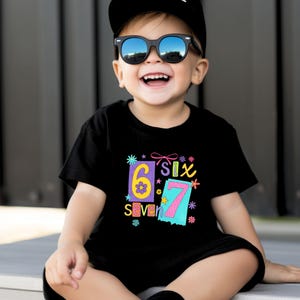 May include: A smiling child wearing sunglasses and a black t-shirt with the numbers "6" and "7" in colorful designs. The shirt also has the word "Six" and "Seven" printed on it. The child is also wearing a black cap.