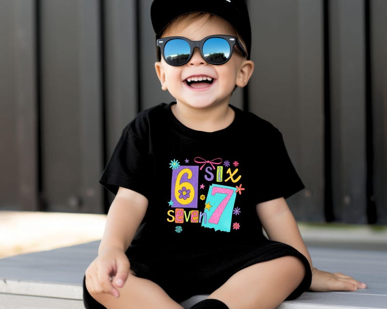 May include: A smiling child wearing sunglasses and a black t-shirt with the numbers "6" and "7" in colorful designs. The shirt also has the word "Six" and "Seven" printed on it. The child is also wearing a black cap.