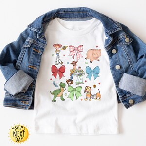 May include: White t-shirt with a colorful design featuring characters from the Toy Story movie franchise. The design includes Woody, Buzz Lightyear, Rex, and other characters. The characters are surrounded by bows and other decorative elements.