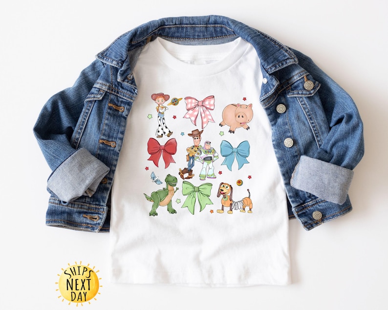 May include: White t-shirt with a colorful design featuring characters from the Toy Story movie franchise. The design includes Woody, Buzz Lightyear, Rex, and other characters. The characters are surrounded by bows and other decorative elements.