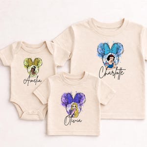May include: A cream-colored baby bodysuit and two cream-colored t-shirts featuring watercolor-style Disney princess graphics with mouse ears. The bodysuit displays Amelia with yellow ears, while the shirts show Olivia with purple ears and Charlotte with blue ears.