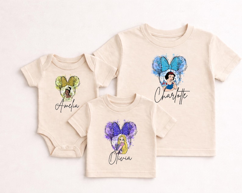 May include: A cream-colored baby bodysuit and two cream-colored t-shirts featuring watercolor-style Disney princess graphics with mouse ears. The bodysuit displays Amelia with yellow ears, while the shirts show Olivia with purple ears and Charlotte with blue ears.