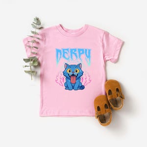 May include: A pink t-shirt featuring the word "DERPY" in blue, and a cartoon blue cat with its tongue sticking out. Two brown leather baby shoes are placed beside the shirt.