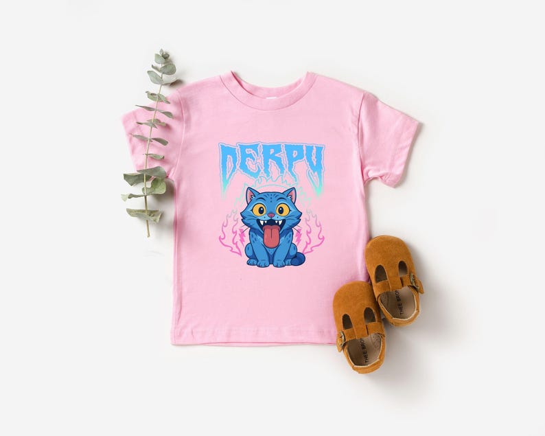 May include: A pink t-shirt featuring the word "DERPY" in blue, and a cartoon blue cat with its tongue sticking out. Two brown leather baby shoes are placed beside the shirt.