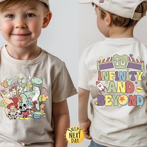 May include: A light beige t-shirt featuring a graphic print of Toy Story characters, including Woody, Buzz Lightyear, Rex, and a dinosaur. The back of the shirt displays the text "To Infinity and Beyond" in colourful letters.