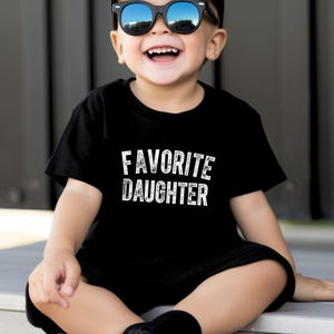 May include: A black t-shirt with the words "FAVORITE DAUGHTER" in white lettering. The t-shirt is being worn by a child who is smiling and wearing sunglasses. The shirt is a crew neck style.