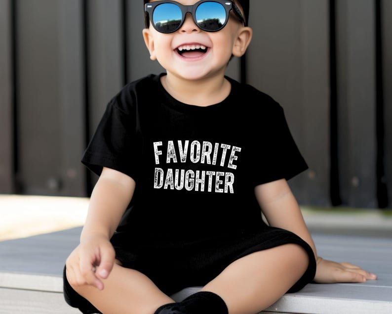 May include: A black t-shirt with the words "FAVORITE DAUGHTER" in white lettering. The t-shirt is being worn by a child who is smiling and wearing sunglasses. The shirt is a crew neck style.