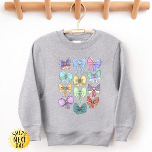May include: A gray sweatshirt with a colorful bow pattern. The bows are in various colors, including pink, blue, yellow, and green. The sweatshirt has a crew neck and long sleeves.