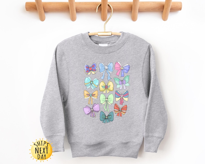 May include: A gray sweatshirt with a colorful bow pattern. The bows are in various colors, including pink, blue, yellow, and green. The sweatshirt has a crew neck and long sleeves.