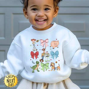 May include: White sweatshirt with a colorful graphic print of characters from the Toy Story movie franchise. The characters are surrounded by bows and hearts.