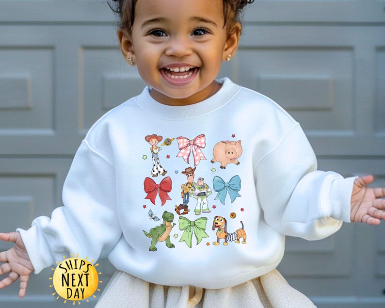 May include: White sweatshirt with a colorful graphic print of characters from the Toy Story movie franchise. The characters are surrounded by bows and hearts.