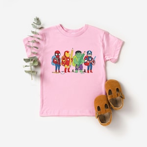 May include: A light pink t-shirt featuring a cartoon illustration of several superheroes, including Spider-Man, Iron Man, Hulk, and Captain America, each holding school supplies. Two brown leather sandals are placed near the shirt.