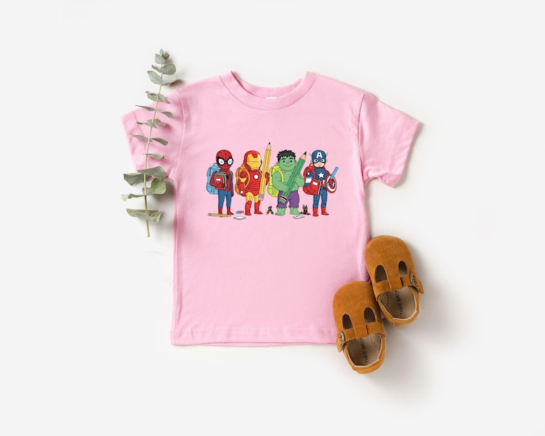 May include: A light pink t-shirt featuring a cartoon illustration of several superheroes, including Spider-Man, Iron Man, Hulk, and Captain America, each holding school supplies. Two brown leather sandals are placed near the shirt.