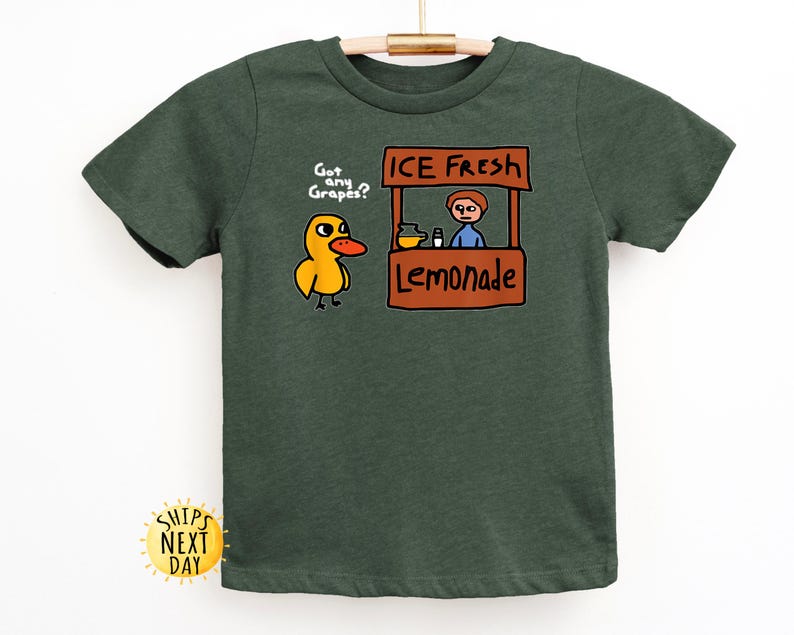 May include: Dark green t-shirt featuring a cartoon duck asking "Got any Grapes?" next to a lemonade stand with the text "ICE FRESH Lemonade." The shirt is made of a soft material and is suitable for children.