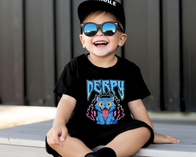 May include: Black t-shirt featuring the word "DERPY" in blue, and a cartoon blue cat with its tongue out, surrounded by pink and purple flames. The child is wearing a black cap and sunglasses.