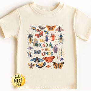May include: Cream-colored t-shirt featuring a colorful insect and butterfly graphic. The design includes various bugs and the text "BE KIND TO ALL KINDS". The shirt is made of soft material.