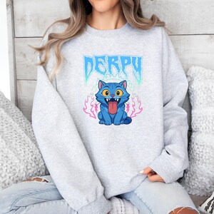 May include: A light grey sweatshirt with a blue cartoon cat graphic, tongue sticking out, and flames. The word "DERPY" is above the cat in a stylized font with blue and teal accents.