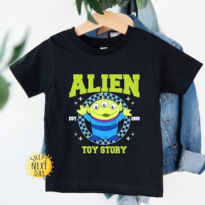 Puede incluir: Camiseta negra con un personaje extraterrestre verde de Toy Story. La camiseta tiene el texto "Alien" en un arco amarillo sobre el personaje y "Toy Story" debajo del personaje. El texto "Est. 1999" est&aacute; en blanco debajo del arco.