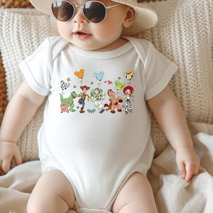 May include: White baby onesie featuring a colorful graphic of characters from a popular animated movie. The design includes a green dinosaur, a cowboy, a space ranger, a horse, and a cowgirl. The onesie is short-sleeved and has a round neckline.