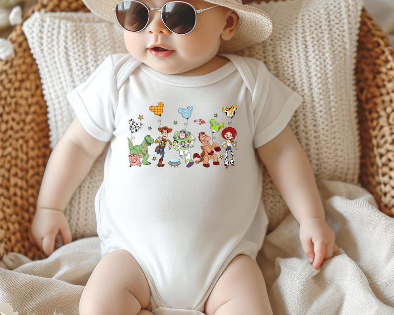 May include: White baby onesie featuring a colorful graphic of characters from a popular animated movie. The design includes a green dinosaur, a cowboy, a space ranger, a horse, and a cowgirl. The onesie is short-sleeved and has a round neckline.