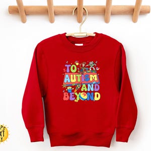 K&ouml;nnte beinhalten: Ein rotes Sweatshirt mit dem Text "TO AUTISM AND BEYOND" in bunten Buchstaben. Das Design beinhaltet Zeichentrickfiguren und eine Rakete. Das Sweatshirt h&auml;ngt an einem Holzb&uuml;gel.