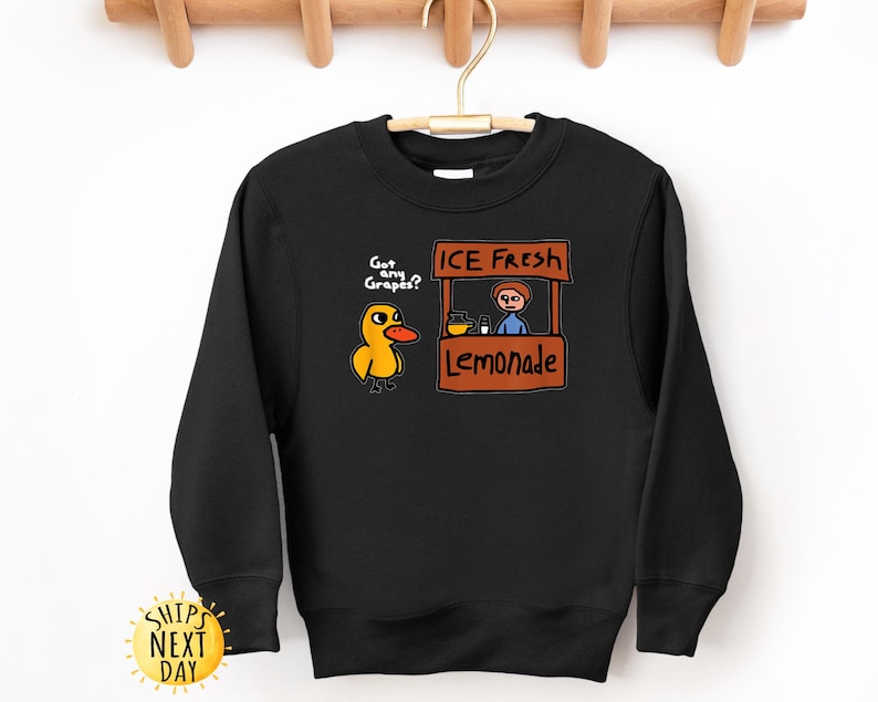 May include: Black sweatshirt with a cartoon duck asking "Got any Grapes?" and a lemonade stand graphic that says "ICE FRESH Lemonade." The sweatshirt has a crew neck and long sleeves. The design is in yellow, orange, brown, and blue.