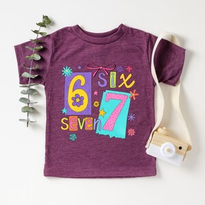 May include: A purple t-shirt with the numbers six and seven in a colorful design. The number six is yellow with a flower, and the number seven is pink. The word "seven" is written in yellow. The shirt has a round neck.