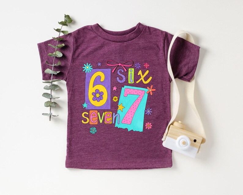 May include: A purple t-shirt with the numbers six and seven in a colorful design. The number six is yellow with a flower, and the number seven is pink. The word "seven" is written in yellow. The shirt has a round neck.