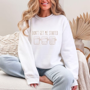 May include: White crew neck sweatshirt with the phrase "DON'T GET ME STARTED" printed in brown. Below the text are three brown jar illustrations. The sweatshirt is a casual, comfortable style.