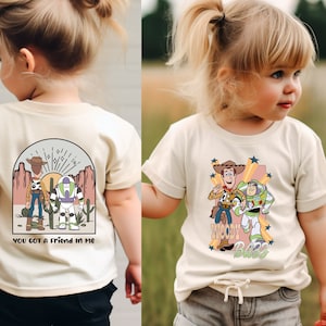 K&ouml;nnte beinhalten: Ein hellbeiges T-Shirt mit einem Grafikdruck von Woody und Buzz Lightyear aus Toy Story. Der Text "You got a friend in me" ist auf der R&uuml;ckseite des Shirts gedruckt.