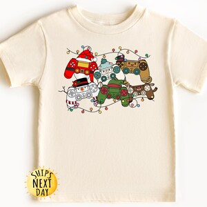 May include: A cream-colored t-shirt with a Christmas-themed design featuring multiple video game controllers decorated as festive characters. The controllers are surrounded by a string of lights.