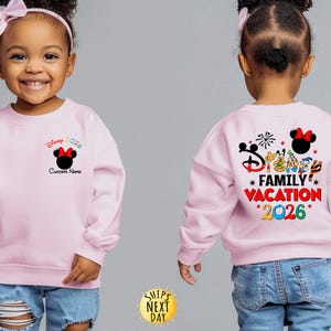 May include: A light pink sweatshirt with a Minnie Mouse graphic and the text "Disney 2026" on the front. The back of the sweatshirt features the text "Disney Family Vacation 2026" with various Disney character illustrations.