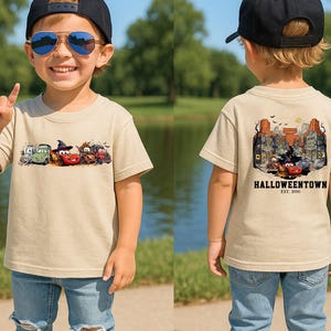 K&ouml;nnte beinhalten: Beiges kurz&auml;rmeliges T-Shirt mit einem Halloween-Motiv auf Vorder- und R&uuml;ckseite. Die Vorderseite zeigt Cartoon-Autos in Halloween-Kost&uuml;men. Die R&uuml;ckseite zeigt eine Stadtlandschaft mit dem Text "Halloweentown Est. 2006".