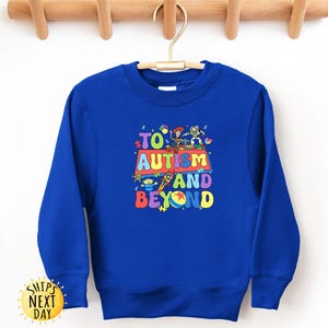 K&ouml;nnte beinhalten: Ein k&ouml;nigsblaues Sweatshirt mit dem Text "TO AUTISM AND BEYOND" in farbenfrohen, verspielten Buchstaben. Das Design beinhaltet Zeichentrickfiguren. Das Sweatshirt hat einen Rundhalsausschnitt und lange &Auml;rmel.