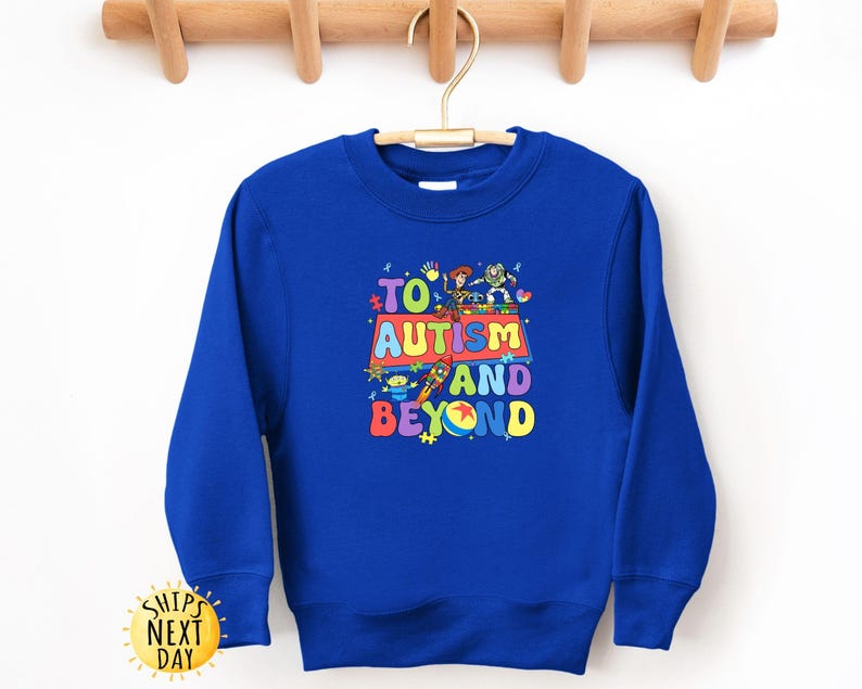 K&ouml;nnte beinhalten: Ein k&ouml;nigsblaues Sweatshirt mit dem Text "TO AUTISM AND BEYOND" in farbenfrohen, verspielten Buchstaben. Das Design beinhaltet Zeichentrickfiguren. Das Sweatshirt hat einen Rundhalsausschnitt und lange &Auml;rmel.