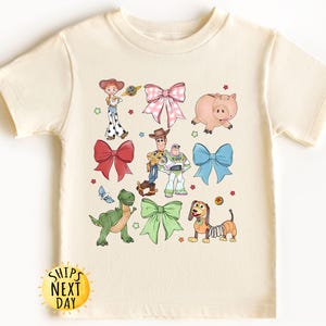 May include: A cream-colored t-shirt with a colorful print of Toy Story characters, including Woody, Buzz Lightyear, Jessie, Rex, and Hamm. The characters are surrounded by bows in various colors. The shirt has a crew neck and short sleeves.