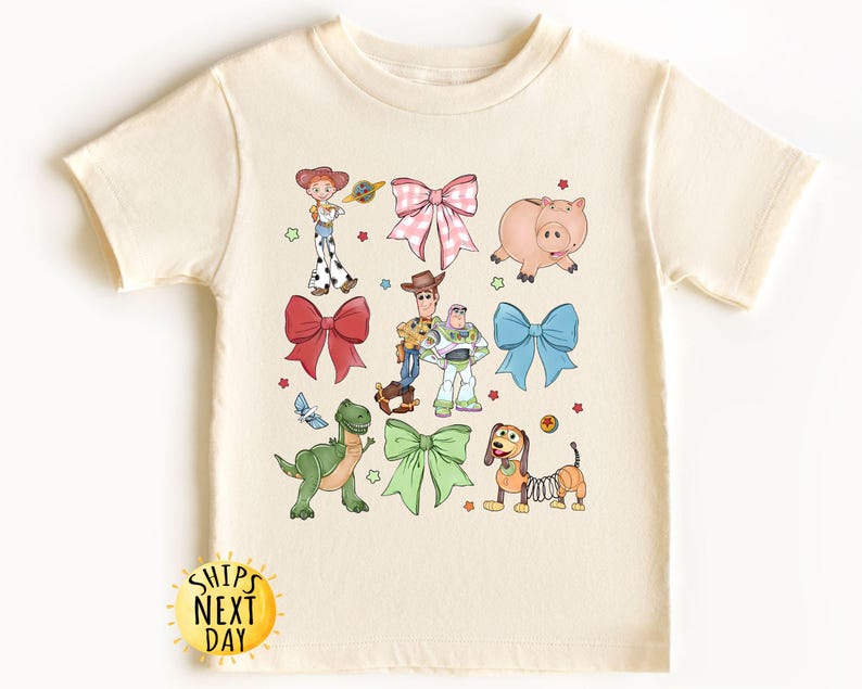 May include: A cream-colored t-shirt with a colorful print of Toy Story characters, including Woody, Buzz Lightyear, Jessie, Rex, and Hamm. The characters are surrounded by bows in various colors. The shirt has a crew neck and short sleeves.