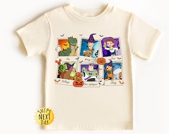 May include: Cream-colored t-shirt featuring a Halloween-themed graphic. The design includes six framed images of cartoon characters dressed in Halloween costumes, with text labels below each image. The shirt has short sleeves and a crew neck.