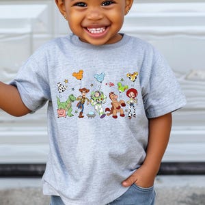May include: A gray t-shirt featuring a colorful graphic of characters from a popular animated movie. The design includes a dinosaur, a pig, a cowboy, a space ranger, a horse, and a cowgirl. The shirt is being worn by a child.