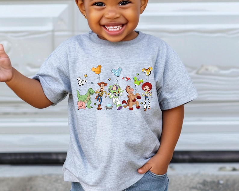 May include: A gray t-shirt featuring a colorful graphic of characters from a popular animated movie. The design includes a dinosaur, a pig, a cowboy, a space ranger, a horse, and a cowgirl. The shirt is being worn by a child.