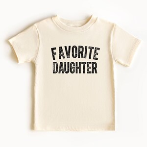 May include: A cream-coloured t-shirt featuring the text "FAVORITE DAUGHTER" in a distressed black font. The shirt has short sleeves and a crew neck. The text is centrally positioned on the front of the garment.