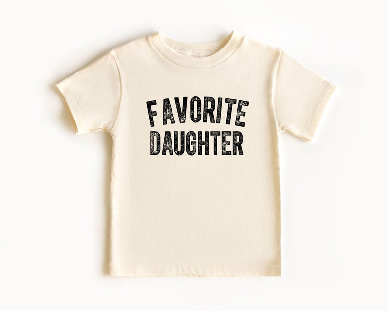 May include: A cream-coloured t-shirt featuring the text "FAVORITE DAUGHTER" in a distressed black font. The shirt has short sleeves and a crew neck. The text is centrally positioned on the front of the garment.