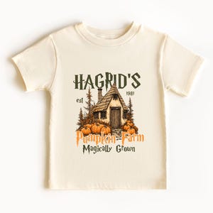 May include: Cream-colored t-shirt featuring a graphic of a cottage with pumpkins and the text "Hagrid's Pumpkin Farm Magically Grown." The design includes the year "1981" and is printed in shades of brown and orange.