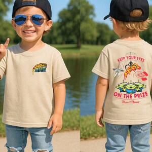 May include: Tan t-shirt featuring a pizza slice graphic on the front and the text "Keep Your Eyes on the Prize" with alien characters on the back. The shirt is worn by a child wearing a black cap and blue jeans.