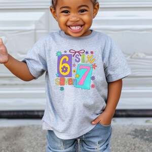 May include: A gray t-shirt with the numbers "6" and "7" in colorful designs. The word "seven" is below the number "6". The shirt is worn by a young child.